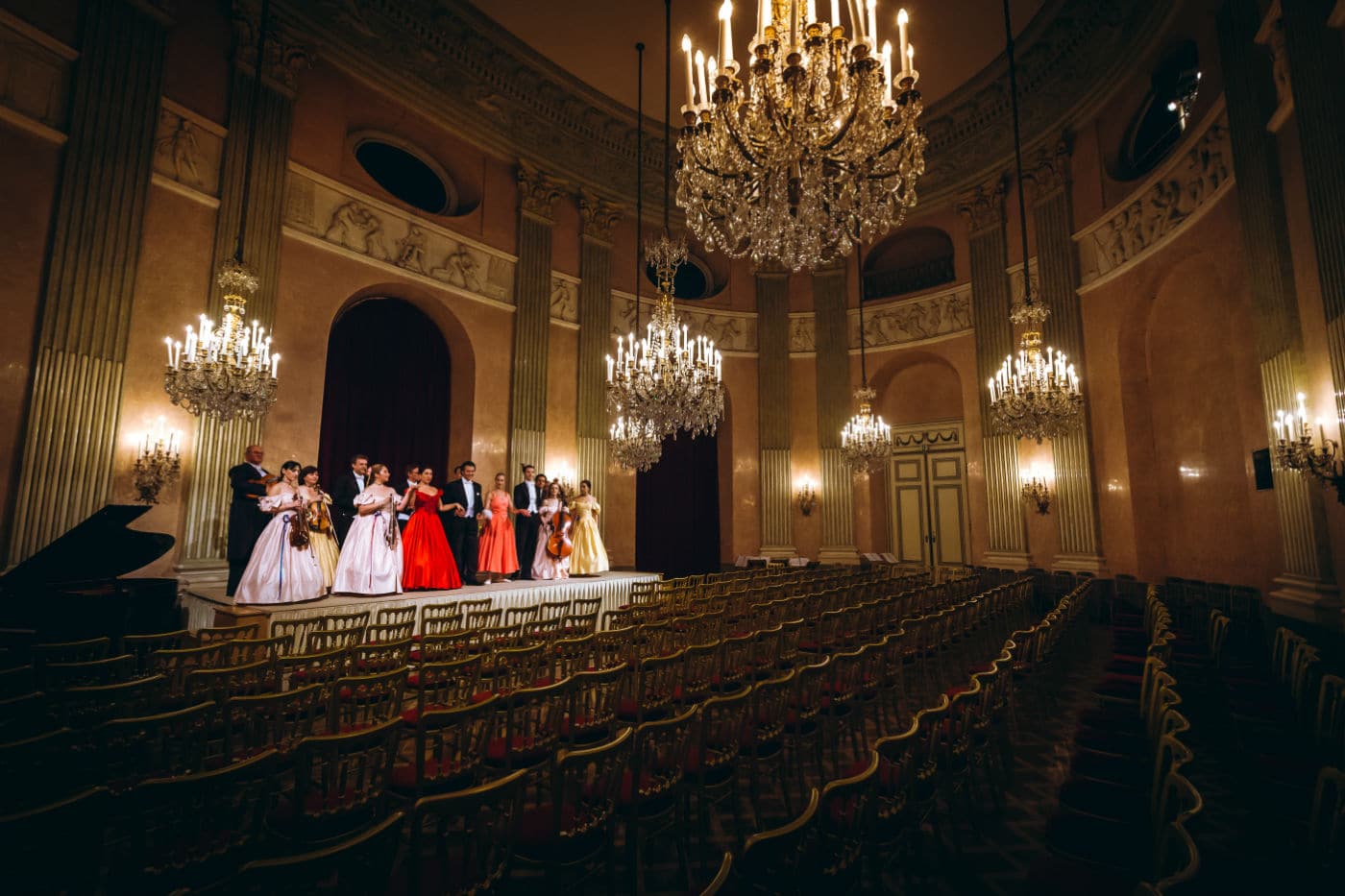 Sala da concerto elegante con sedie dorate e lampadari accesi, orchestra in abiti da gala sul palco pronta per la performance.