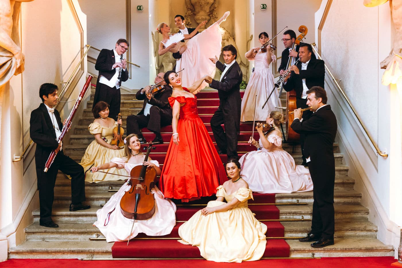 Gruppo di musicisti e ballerini in costumi d’epoca posizionati su una scalinata interna, con strumenti musicali e gesti scenici.