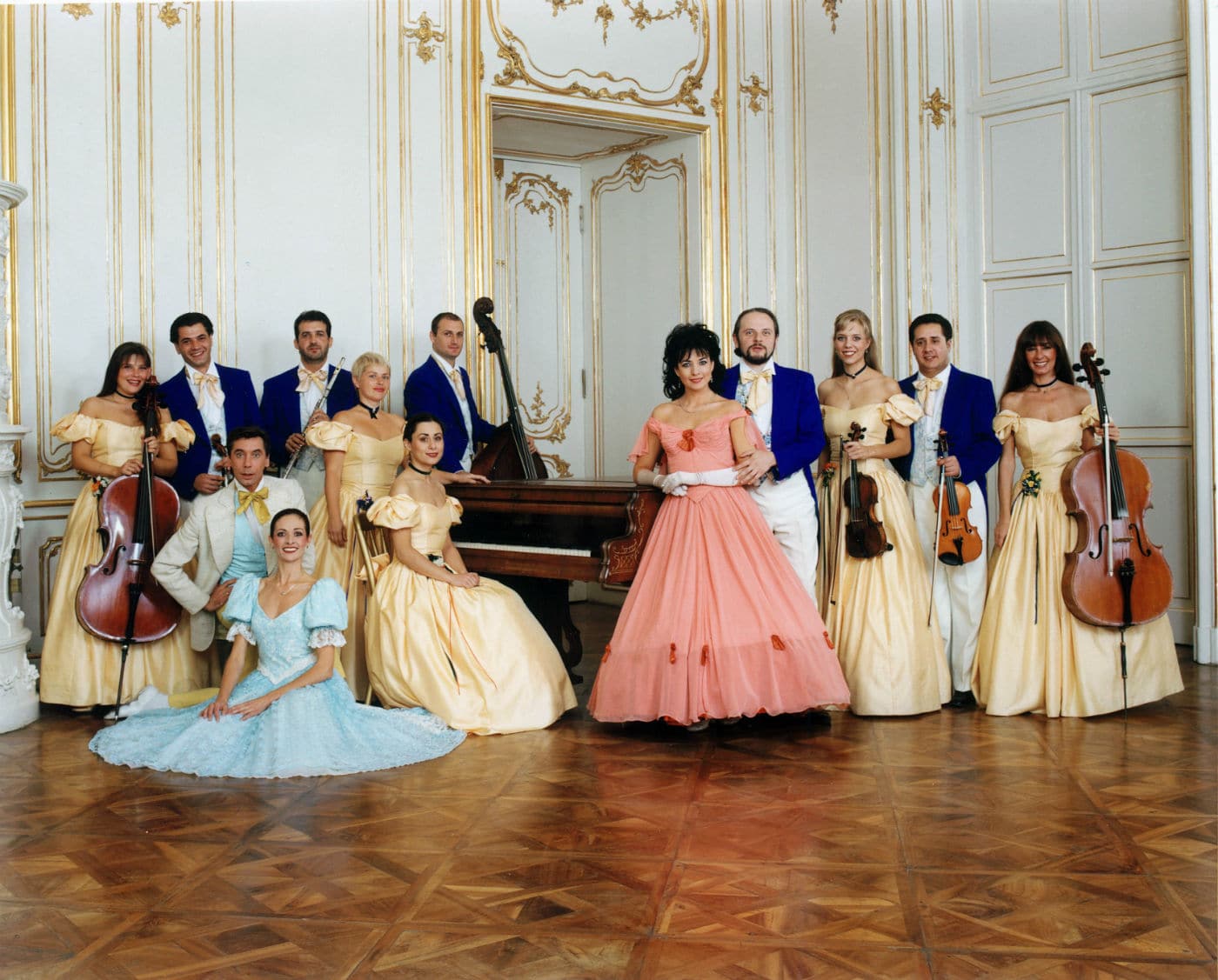 Musicisti e ballerini in costumi d’epoca posano davanti a un pianoforte in una sala decorata in stile barocco.