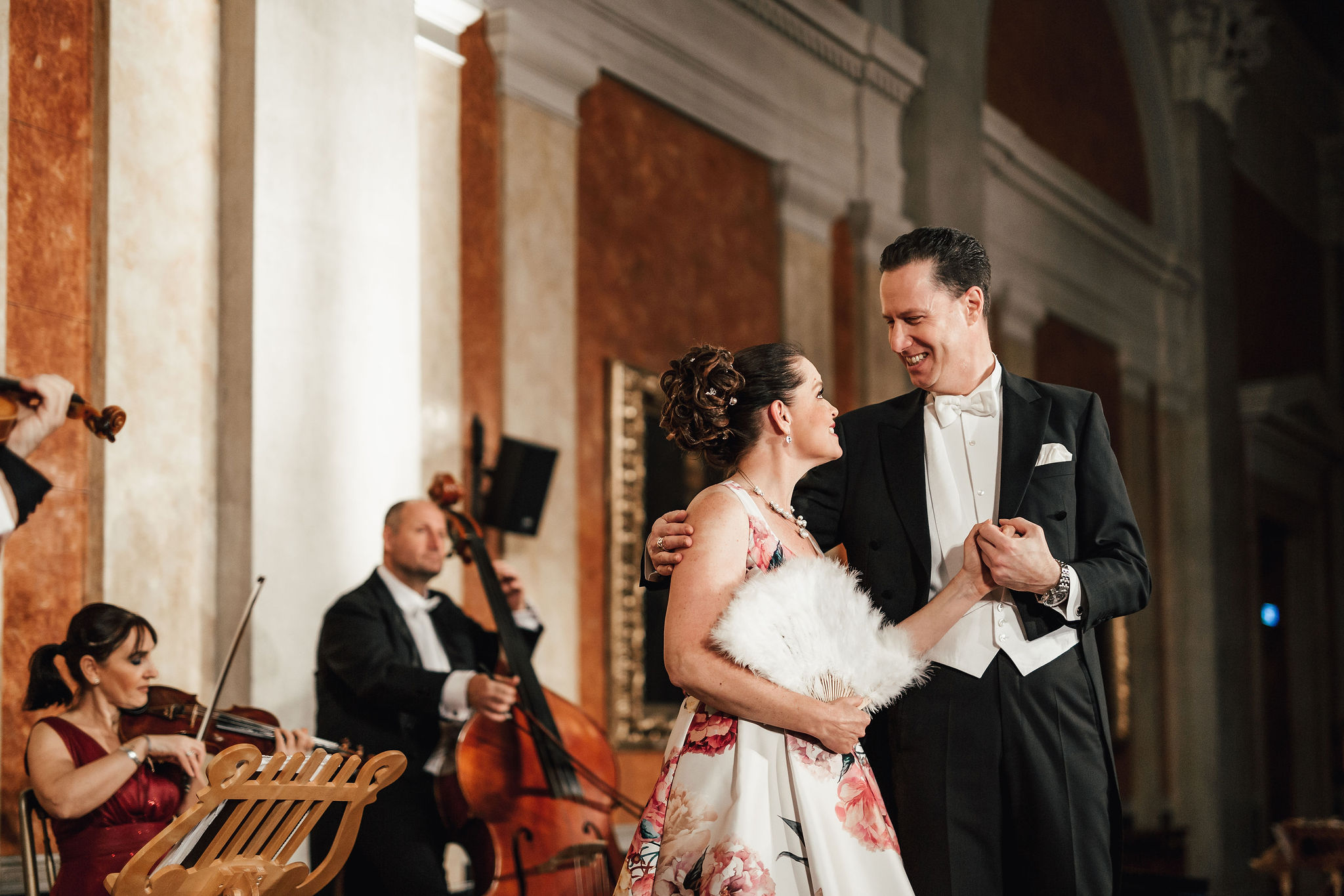 Coppia in abiti da sera balla il valzer, accompagnata da musicisti in sala da concerto storica.