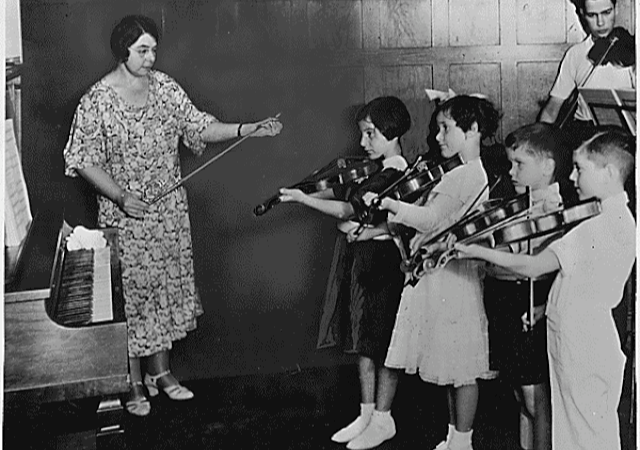 Historische Geigen-Unterrichtsstunde für Kinder Historische Schwarz-Weiß-Aufnahme einer Geigen-Unterrichtsstunde. Eine Lehrerin steht vor einer Gruppe von Kindern und unterrichtet sie, während die Kinder ihre Geigen in Spielposition halten.