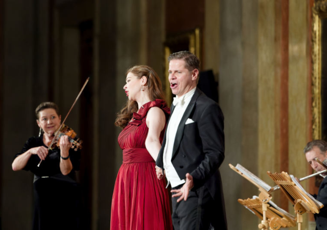 2 Ein Mann im Frack und eine Frau im roten Abendkleid singen leidenschaftlich auf einer Bühne, begleitet von einer lächelnden Geigerin und weiteren Musikern eines klassischen Orchesters im Hintergrund.