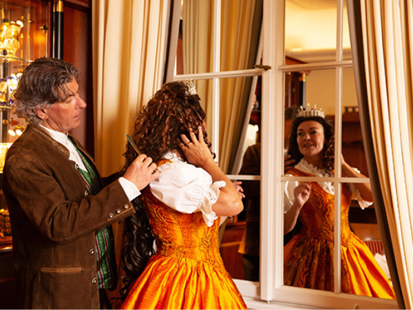 Monika Mosser und Schauspielkollege bereiten sich auf die Szene vor Monika Mosser in orangefarbenem historischen Kleid steht vor einem Fenster und richtet ihr Haar, während ein männlicher Schauspieler hinter ihr steht und sie berührt.
