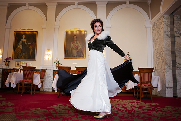 Monika Mosser dreht sich elegant in einem weißen und schwarzen Ballkleid im festlich eingerichteten Saal mit Gemälden an den Wänden und roten Teppichen.